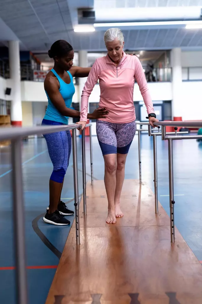 A hospital rehabilitation room bathed in soft, natural lighting. In the foreground, a patient wearing physical therapy attire steadily performs knee extension exercises on a cushioned mat, their determination etched on their face. In the middle ground, a physical therapist observes closely, providing gentle guidance and encouragement. The background reveals various rehabilitation equipment, including parallel bars, exercise balls, and a wall-mounted series of mirrors, creating a serene, clinical atmosphere conducive to the recovery process. The overall scene conveys a sense of progress, resilience, and the collaborative effort between the patient and healthcare provider towards successful bilateral knee replacement rehabilitation. A hospital rehabilitation room bathed in soft, natural lighting. In the foreground, a patient wearing physical therapy attire steadily performs knee extension exercises on a cushioned mat, their determination etched on their face. In the middle ground, a physical therapist observes closely, providing gentle guidance and encouragement. The background reveals various rehabilitation equipment, including parallel bars, exercise balls, and a wall-mounted series of mirrors, creating a serene, clinical atmosphere conducive to the recovery process. The overall scene conveys a sense of progress, resilience, and the collaborative effort between the patient and healthcare provider towards successful bilateral knee replacement rehabilitation.