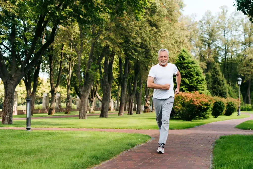 A middle-aged person in athletic wear carefully and cautiously running on a paved trail, their face determined yet slightly pained. The runner's posture is upright and their gait is measured, indicating a sense of controlled motion after recent knee replacement surgery. The scene is captured in a medium shot, with the runner positioned in the center of the frame against a backdrop of lush greenery and a clear blue sky. The lighting is natural and soft, casting a warm, calming glow on the scene. The atmosphere conveys a sense of the runner's journey towards recovery and the medical perspectives on resuming high-impact activities after such a procedure.
