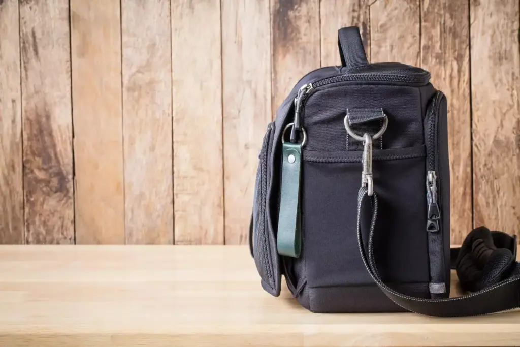 A sleek and functional chemotherapy backpack rests on a wooden table, its straps and pockets neatly arranged. The backpack is a soft gray color, with a subtle textured fabric that conveys a sense of durability and comfort. The design features multiple compartments, allowing for efficient organization of essential items such as medication, water bottles, and personal care products. Warm, diffused lighting from above gently illuminates the scene, creating a calming and reassuring atmosphere. The backpack is positioned in the foreground, with a slightly blurred background that suggests a clean, minimalist space, allowing the focus to remain on the practical yet stylish medical accessory.