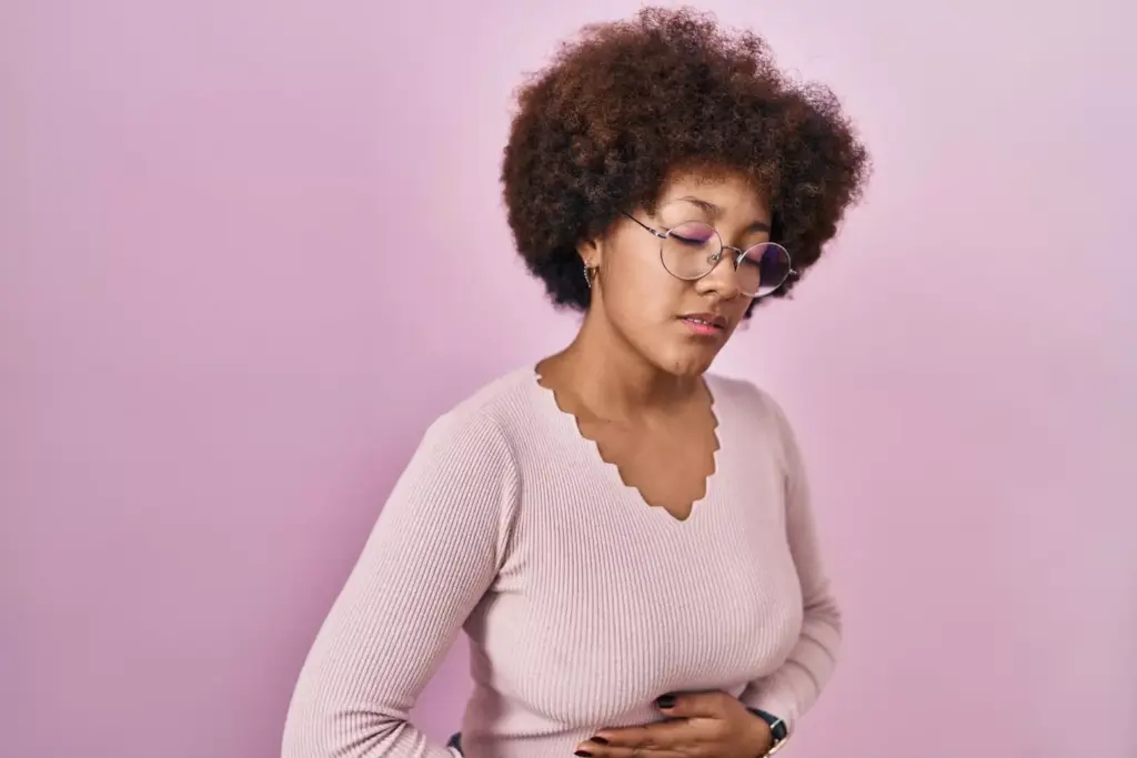 Young african american woman standing over pink background with hand on stomach because indigestion, painful illness feeling unwell. ache concept.